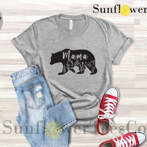 May include: A light grey heather t-shirt with a black silhouette of a mama bear with floral details and the word "Mama" inside the bear. The shirt is laid out on a wooden surface with a pair of blue jeans and red sneakers.