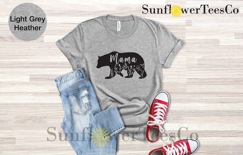 May include: A light grey heather t-shirt with a black silhouette of a mama bear with floral details and the word "Mama" inside the bear. The shirt is laid out on a wooden surface with a pair of blue jeans and red sneakers.
