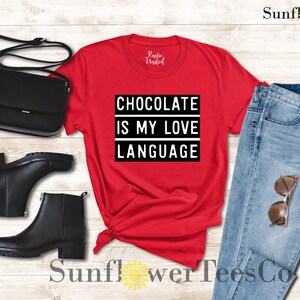 May include: A red t-shirt with the text "CHOCOLATE IS MY LOVE LANGUAGE" in a black and white rectangular design. The image also includes black ankle boots, a black purse, light blue jeans, and sunglasses.