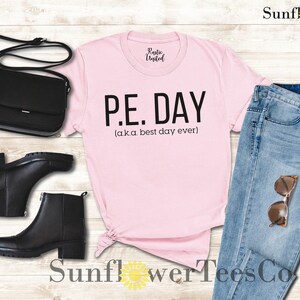Op de afbeelding: Roze T-shirt met zwarte tekst die "P.E. DAY (aka best day ever)" zegt. Het shirt ligt op een houten oppervlak met een zwarte tas, zwarte laarzen, een blauwe jeans en een zonnebril.