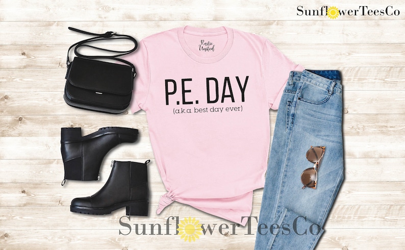 Op de afbeelding: Roze T-shirt met zwarte tekst die "P.E. DAY (aka best day ever)" zegt. Het shirt ligt op een houten oppervlak met een zwarte tas, zwarte laarzen, een blauwe jeans en een zonnebril.