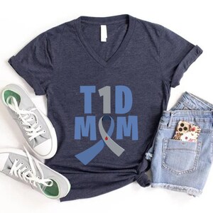 Pu&ograve; includere: Una maglietta blu navy con scollo a V bianco. La maglietta ha un nastro blu e grigio con una goccia di sangue rossa sopra. Il testo "T1D MOM" &egrave; stampato sulla maglietta.