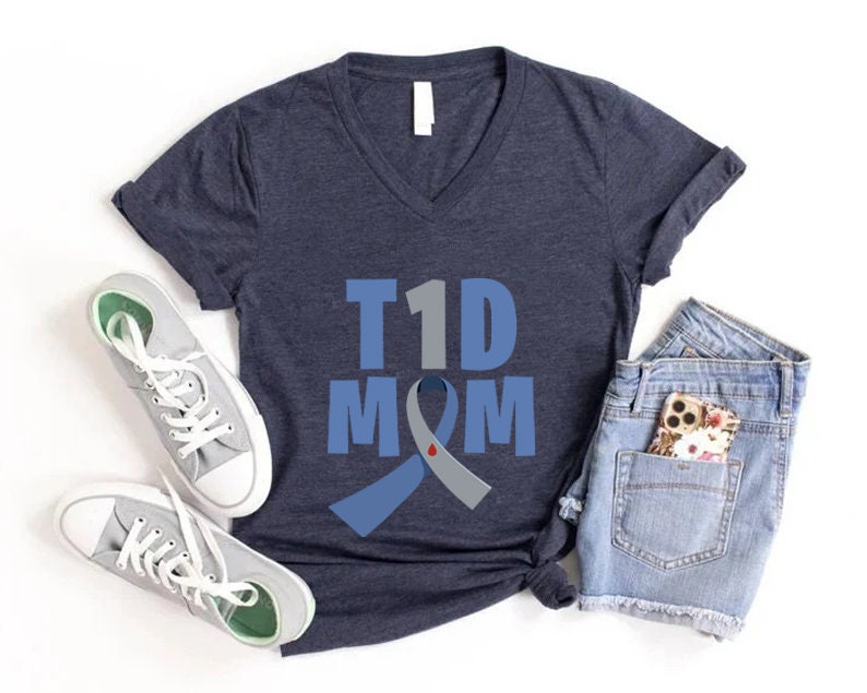 Pu&ograve; includere: Una maglietta blu navy con scollo a V bianco. La maglietta ha un nastro blu e grigio con una goccia di sangue rossa sopra. Il testo "T1D MOM" &egrave; stampato sulla maglietta.
