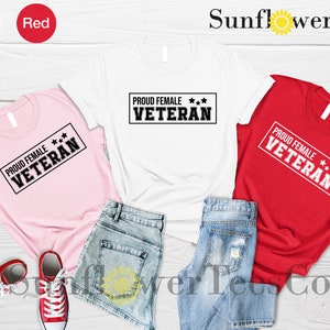 May include: Three t-shirts in pink, white, and red with the text "PROUD FEMALE*** VETERAN" printed on each. The shirts are laid out on a wooden surface with denim shorts.