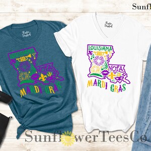 May include: Two t-shirts with a purple and green outline of the state of Louisiana with Mardi Gras themed graphics. The text "Louisiana" and "Mardi Gras" are in yellow. The t-shirts are on a wooden background with a pair of black boots and blue jeans.