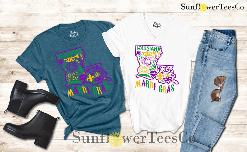 May include: Two t-shirts with a purple and green outline of the state of Louisiana with Mardi Gras themed graphics. The text "Louisiana" and "Mardi Gras" are in yellow. The t-shirts are on a wooden background with a pair of black boots and blue jeans.