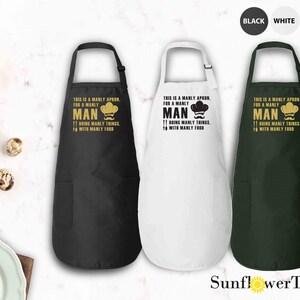 May include: Three black, white, and green aprons with a pocket on the bottom. The aprons have a gold chef hat and mustache graphic with the text "This is a manly apron, for a manly man doing manly things with manly food."