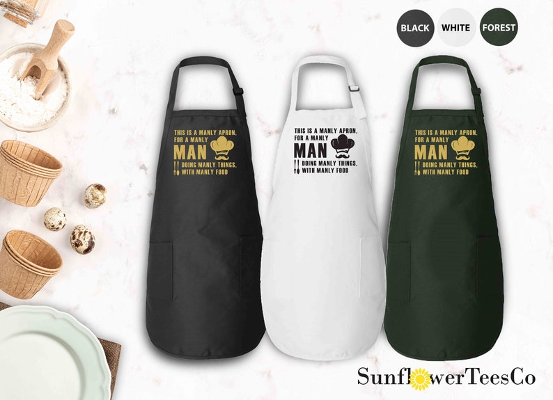 May include: Three black, white, and green aprons with a pocket on the bottom. The aprons have a gold chef hat and mustache graphic with the text "This is a manly apron, for a manly man doing manly things with manly food."