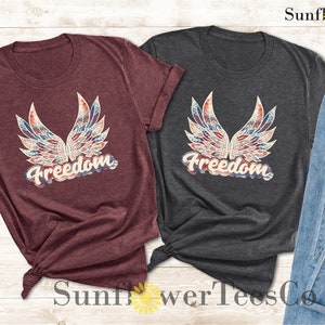 Puede incluir: Dos camisetas con un dise&ntilde;o estampado de alas en rojo, blanco y azul y la palabra "Freedom" en una fuente vintage. Las camisetas son granate y gris oscuro.