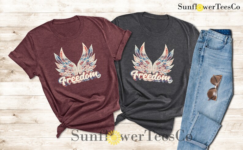 Puede incluir: Dos camisetas con un dise&ntilde;o estampado de alas en rojo, blanco y azul y la palabra "Freedom" en una fuente vintage. Las camisetas son granate y gris oscuro.