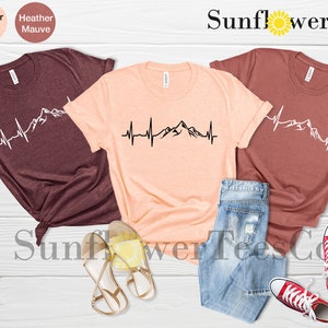 May include: Three t-shirts with a mountain range and heartbeat graphic design. The t-shirts are in heather maroon, heather peach, and heather mauve colors. The text "Sunflower TeesCo" is visible at the top of the image.