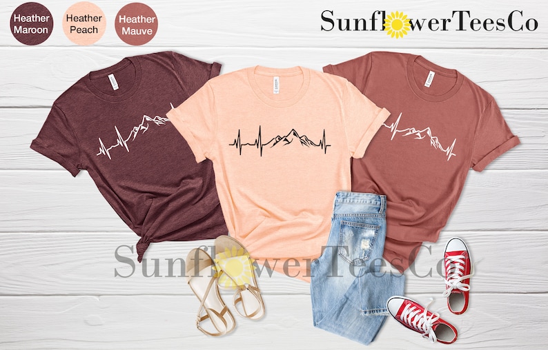 May include: Three t-shirts with a mountain range and heartbeat graphic design. The t-shirts are in heather maroon, heather peach, and heather mauve colors. The text "Sunflower TeesCo" is visible at the top of the image.