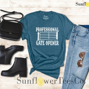 Op de afbeelding: Een tealblauw t-shirt met de tekst "Professional Gate Opener" en een wit silhouet van een poort. Het shirt ligt op een houten achtergrond met een paar zwarte laarzen, een zwarte tas en een blauwe jeans met een zonnebril.