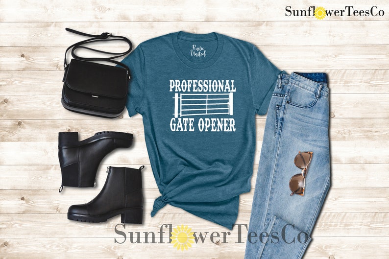 Op de afbeelding: Een tealblauw t-shirt met de tekst "Professional Gate Opener" en een wit silhouet van een poort. Het shirt ligt op een houten achtergrond met een paar zwarte laarzen, een zwarte tas en een blauwe jeans met een zonnebril.