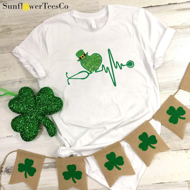 May include: White t-shirt with a green shamrock heart and a stethoscope with a shamrock on the end. The heart has a green leprechaun hat on top. The text "SunflowerTeesCo" is in the top left corner of the image.