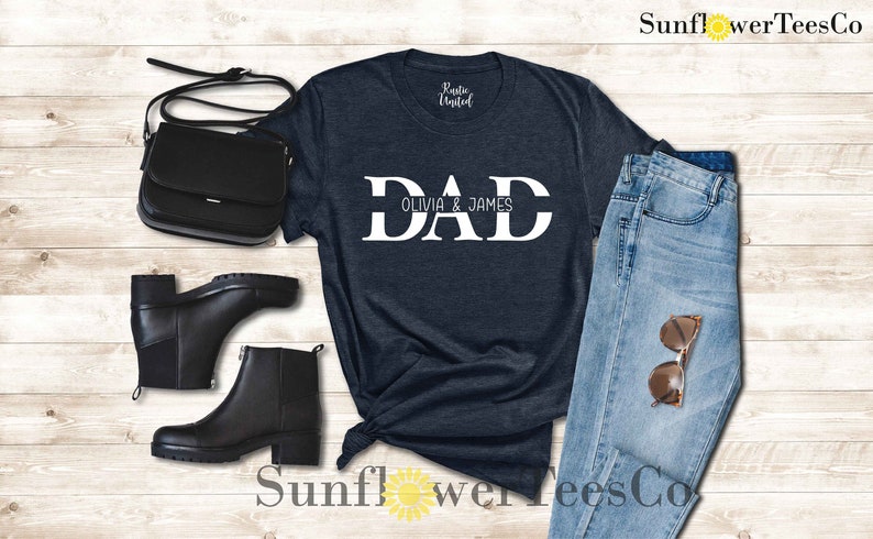 Op de afbeelding: Een marineblauw t-shirt met het woord "DAD" in witte blokletters. De tekst "Olivia & James" is in kleinere witte letters onder het woord "DAD" gedrukt.
