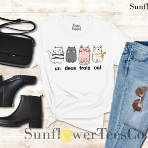 以下が含まれることがあります： 4匹の猫が並んだグラフィックがプリントされた白いTシャツ。猫はそれぞれ違う服を着ていて、表情も違います。「un deux trois cat」という文字が猫の下にプリントされています。