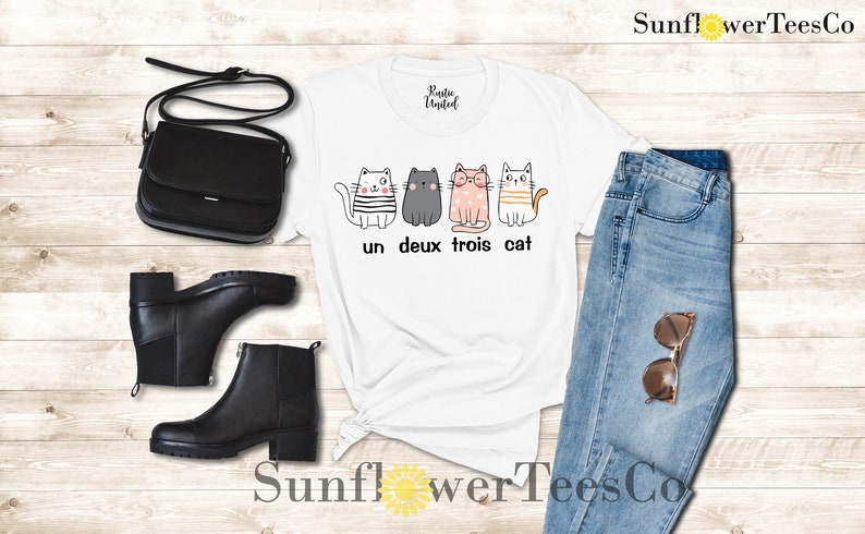 以下が含まれることがあります： 4匹の猫が並んだグラフィックがプリントされた白いTシャツ。猫はそれぞれ違う服を着ていて、表情も違います。「un deux trois cat」という文字が猫の下にプリントされています。
