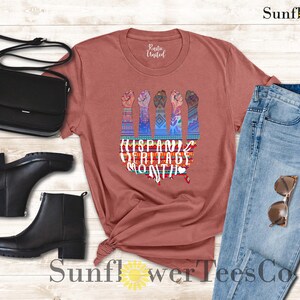 May include: A rust-colored t-shirt with a graphic of five raised fists with colorful, patterned sleeves. The text "Hispanic Heritage Month" is printed below the fists. The t-shirt is displayed on a wooden background with a pair of black boots, a black purse, and blue jeans.