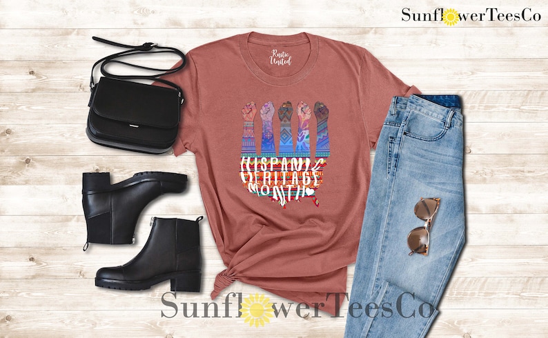 May include: A rust-colored t-shirt with a graphic of five raised fists with colorful, patterned sleeves. The text "Hispanic Heritage Month" is printed below the fists. The t-shirt is displayed on a wooden background with a pair of black boots, a black purse, and blue jeans.