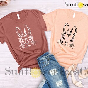 May include: Two t-shirts with a graphic of a bunny wearing glasses. The shirt on the left is a maroon color and the shirt on the right is a peach color. The text "Sunflower TeesCo" is visible in the top right corner of the image.