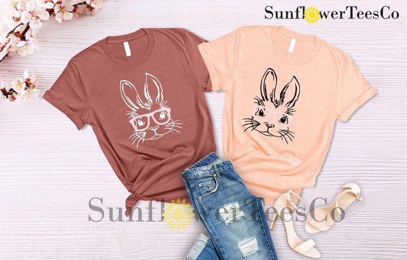 May include: Two t-shirts with a graphic of a bunny wearing glasses. The shirt on the left is a maroon color and the shirt on the right is a peach color. The text "Sunflower TeesCo" is visible in the top right corner of the image.