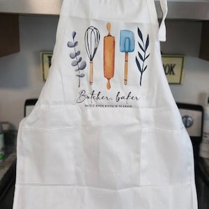 K&ouml;nnte beinhalten: Wei&szlig;er Sch&uuml;rze mit einem Aquarelldesign von K&uuml;chenutensilien, darunter ein Schneebesen, eine Teigrolle und ein Spatel. Der Text "Butcher, baker, NOT CANDLESTICK MAKER" ist darunter gedruckt. Die Sch&uuml;rze hat zwei Vordertaschen.