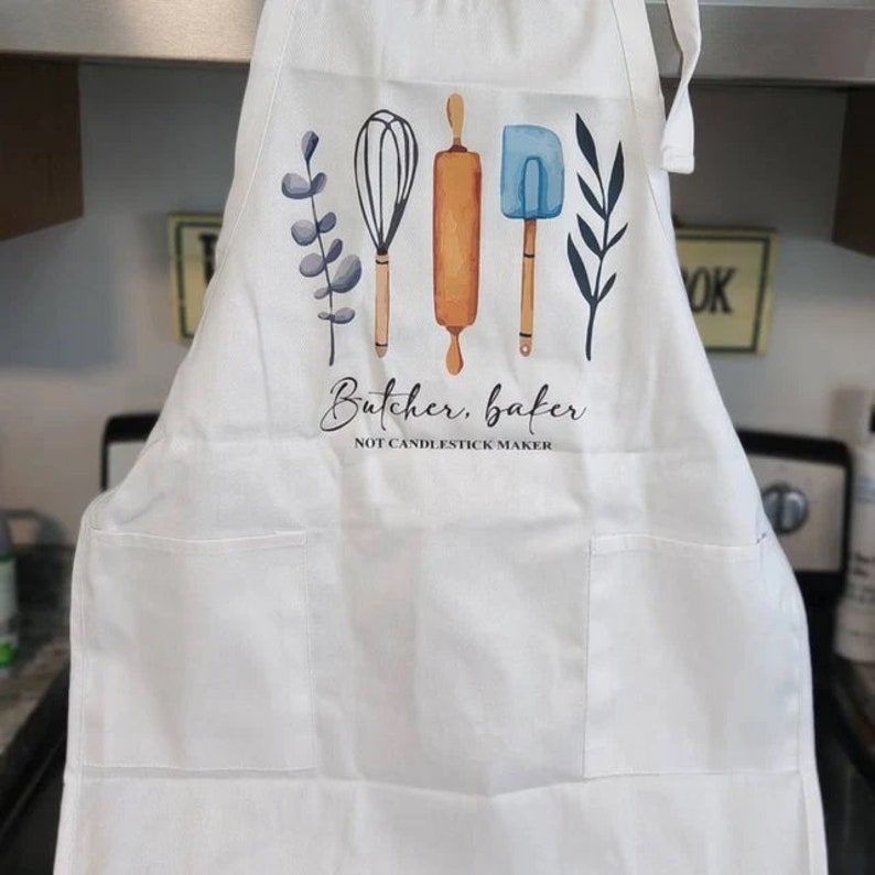 K&ouml;nnte beinhalten: Wei&szlig;er Sch&uuml;rze mit einem Aquarelldesign von K&uuml;chenutensilien, darunter ein Schneebesen, eine Teigrolle und ein Spatel. Der Text "Butcher, baker, NOT CANDLESTICK MAKER" ist darunter gedruckt. Die Sch&uuml;rze hat zwei Vordertaschen.
