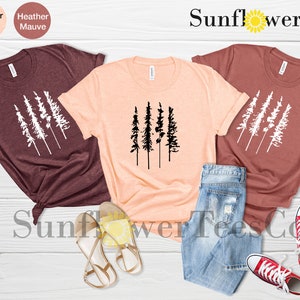 Op de afbeelding: Drie T-shirts met een afbeelding van vijf gestileerde bomen. De shirts zijn in de kleuren heidebruin, perzik en mauve. De afbeelding is wit op de heidebruine en mauve shirts en zwart op het perzik shirt. De shirts liggen op een licht houten oppervlak met een paar sandalen en een spijkerbroek. De tekst "Sunflower TeesCo" is zichtbaar in de rechterbovenhoek van de afbeelding.