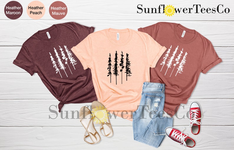 Op de afbeelding: Drie T-shirts met een afbeelding van vijf gestileerde bomen. De shirts zijn in de kleuren heidebruin, perzik en mauve. De afbeelding is wit op de heidebruine en mauve shirts en zwart op het perzik shirt. De shirts liggen op een licht houten oppervlak met een paar sandalen en een spijkerbroek. De tekst "Sunflower TeesCo" is zichtbaar in de rechterbovenhoek van de afbeelding.