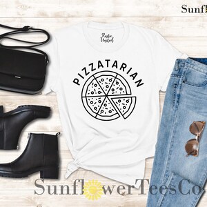 May include: White t-shirt with black text that says "Pizzatarian" and a black and white illustration of a pizza. The shirt is laying on a wooden surface with a pair of black boots, a black purse, and blue jeans with brown sunglasses.