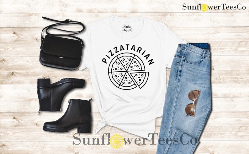 May include: White t-shirt with black text that says "Pizzatarian" and a black and white illustration of a pizza. The shirt is laying on a wooden surface with a pair of black boots, a black purse, and blue jeans with brown sunglasses.