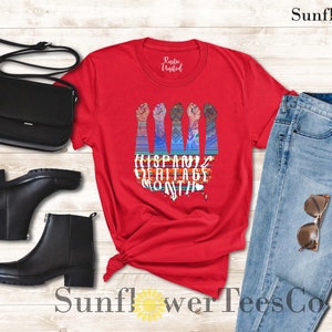 May include: Red t-shirt with a graphic of five raised fists with colorful patterns and the text "Hispanic Heritage Month". The shirt is displayed with a pair of black ankle boots, a black purse, and blue jeans.