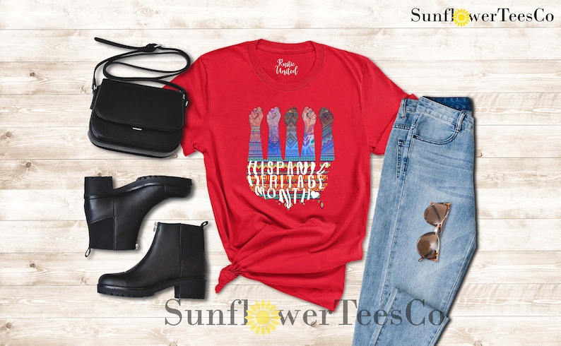May include: Red t-shirt with a graphic of five raised fists with colorful patterns and the text "Hispanic Heritage Month". The shirt is displayed with a pair of black ankle boots, a black purse, and blue jeans.