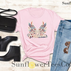 Puede incluir: Camiseta rosa con un gr&aacute;fico de alas y la palabra "Freedom" en estilo vintage. La camiseta se combina con jeans azules, botas negras y un bolso negro.
