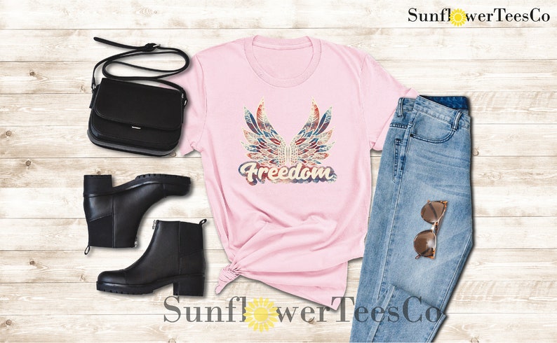 Puede incluir: Camiseta rosa con un gr&aacute;fico de alas y la palabra "Freedom" en estilo vintage. La camiseta se combina con jeans azules, botas negras y un bolso negro.