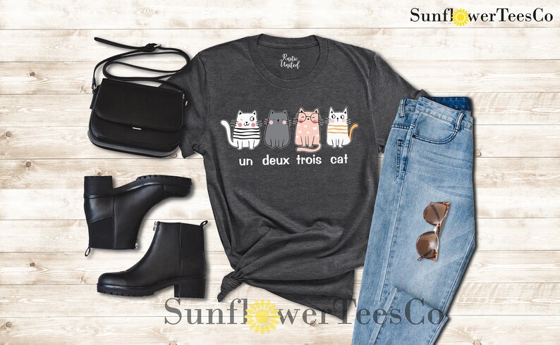 以下が含まれることがあります： 4匹の猫が並んだデザインのダークグレーのTシャツ。猫は白、グレー、ピンク、イエロー。猫の下のテキストは「un deux trois cat」。
