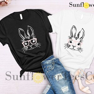 May include: Two t-shirts with a black and white illustration of a bunny wearing pink glasses. The t-shirts are on a white wooden surface with a pair of beige high heels and blue jeans.