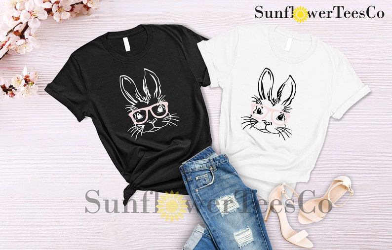 May include: Two t-shirts with a black and white illustration of a bunny wearing pink glasses. The t-shirts are on a white wooden surface with a pair of beige high heels and blue jeans.