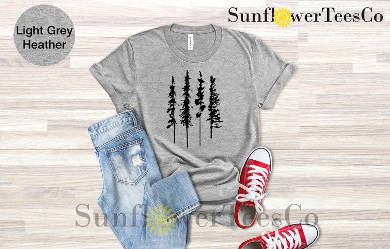 Op de afbeelding: Een lichtgrijs heather t-shirt met een zwart silhouet van vier dennenbomen. Het shirt ligt plat op een houten oppervlak met een blauwe spijkerbroek en rode sneakers.
