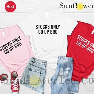 May include: Three t-shirts with the text "Stocks Only Go Up Bro" in black on a pink, white, and red shirt. The shirts are laid out on a wooden surface with denim shorts.