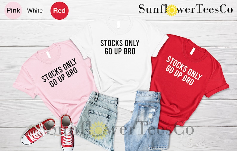 May include: Three t-shirts with the text "Stocks Only Go Up Bro" in black on a pink, white, and red shirt. The shirts are laid out on a wooden surface with denim shorts.