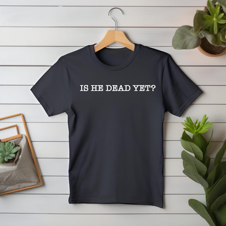K&ouml;nnte beinhalten: Ein dunkelblaues T-Shirt mit dem Aufdruck "IS HE DEAD YET?" in Wei&szlig; auf der Vorderseite.