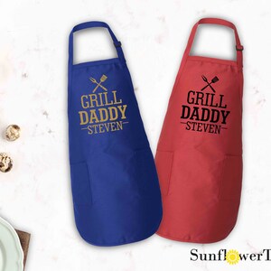 May include: Two aprons with adjustable neck straps and a large pocket. The aprons are blue and red. The text on the aprons reads "Grill Daddy - Steven".