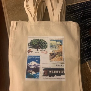 K&ouml;nnte beinhalten: Cremefarbene Tragetaschen mit angebrachten Griffen. Jede Tasche zeigt eine Collage aus alten Briefmarken. Die Briefmarken zeigen eine Wei&szlig;eiche, eine Berglandschaft, eine Strandszene und eine Stra&szlig;enbahn. Die Briefmarken tragen den Text "USA" und verschiedene Nennwerte.