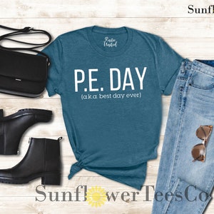 Op de afbeelding: Een tealblauw t-shirt met witte tekst die "P.E. DAY (a.k.a best day ever)" luidt. Het shirt is gestyled met een blauwe jeans, zwarte laarzen en een zwarte handtas.