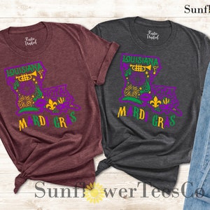 May include: Two t-shirts with a Mardi Gras design featuring a purple, green, and gold Louisiana outline with a trumpet, fleur-de-lis, and a mask. The shirts are maroon and dark grey.