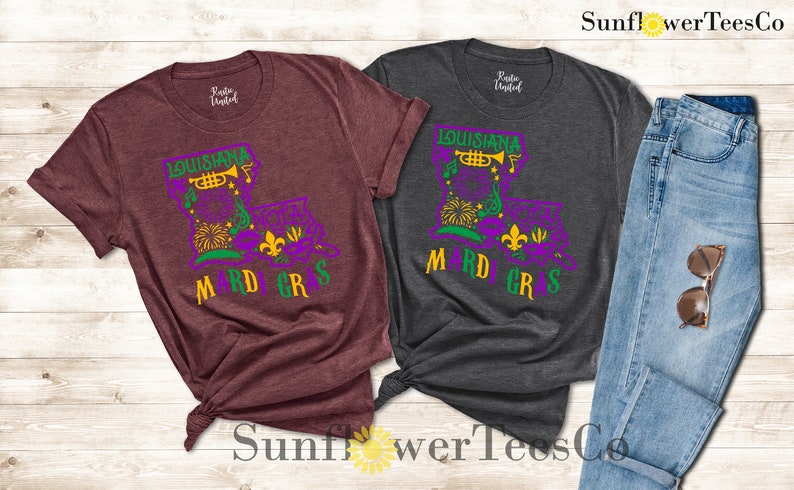 May include: Two t-shirts with a Mardi Gras design featuring a purple, green, and gold Louisiana outline with a trumpet, fleur-de-lis, and a mask. The shirts are maroon and dark grey.