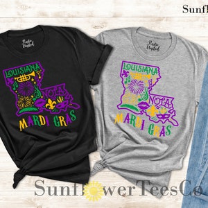 May include: Two t-shirts with a graphic design of the state of Louisiana with the words "Louisiana Mardi Gras" and "NOLA" on them. The shirts are black and grey.