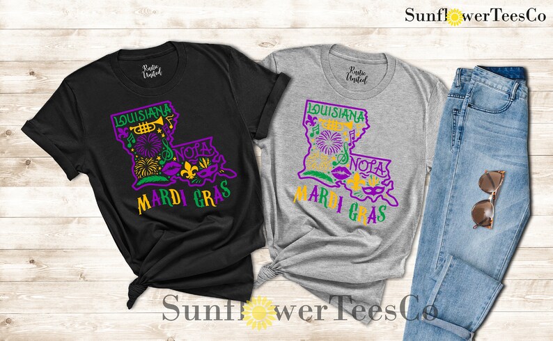 May include: Two t-shirts with a graphic design of the state of Louisiana with the words "Louisiana Mardi Gras" and "NOLA" on them. The shirts are black and grey.